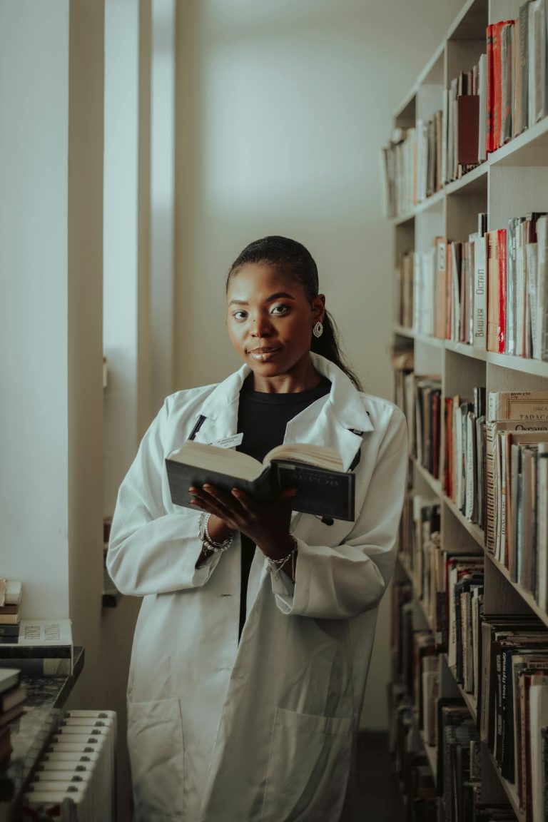 A focused woman in a lab coat reading a book in a library, showcasing education and learning.